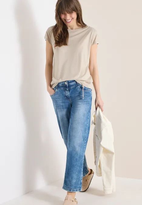 Eine Frau in beigem Oberteil, blauer Jeans, hält weiße Jacke, steht vor weißem Hintergrund.
